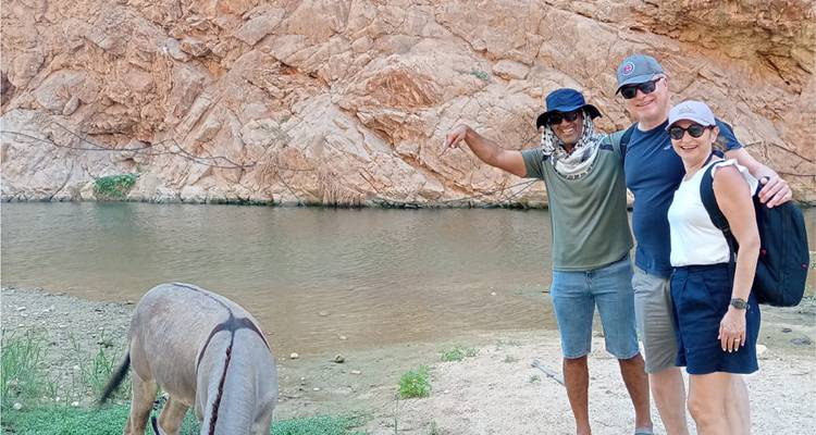 Un guide et deux touristes souriants à côté d'un âne qui boit l'eau calme du canyon.