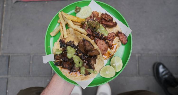 Vue aérienne de deux tacos de rue garnis de viande grillée, de frites, de salsa et de quartiers de citron vert sur une assiette vert vif tenue au-dessus du pavé.