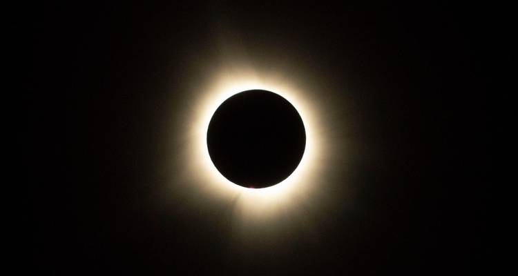 Totale Sonnenfinsternis mit dunkler Mondsscheibe, umgeben von heller Korona vor schwarzem Himmel