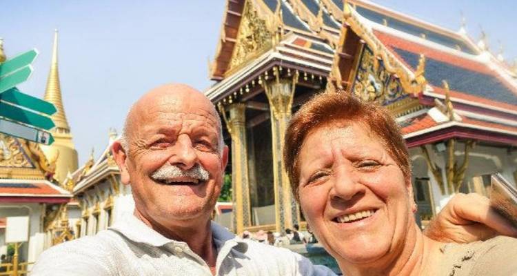 Un couple de personnes âgées souriant prend un selfie devant des bâtiments de temple thaïlandais ornés lors d'une belle journée ensoleillée.