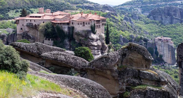 Een afgelegen klooster in Meteora kroont een dramatische rotspilaer boven een groen dal en rollende heuvels.