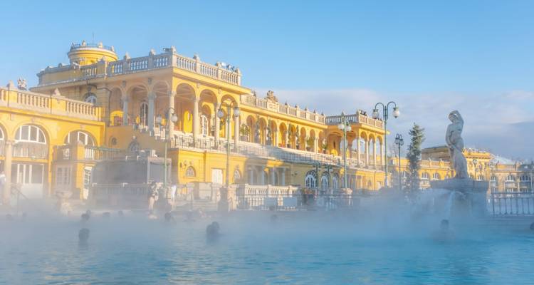 La vapeur s'élève des piscines extérieures turquoise des bains thermaux Széchenyi aux colonnes jaunes sous un ciel bleu.