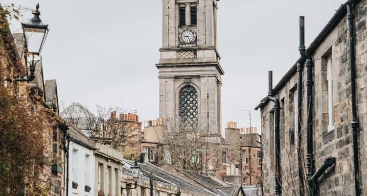 La tour de l'horloge d'une église en pierre domine une ruelle pavée étroite bordée de petits chalets pittoresques par un jour nuageux.