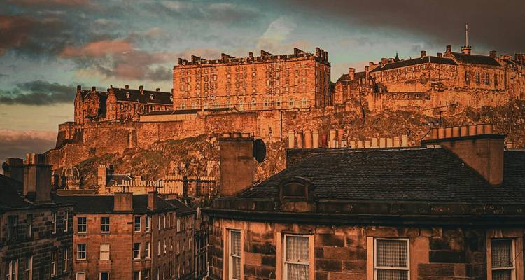 Le Château d'Édimbourg brille dans la lumière chaude du coucher de soleil au-dessus de rangées de toits d'immeubles historiques.