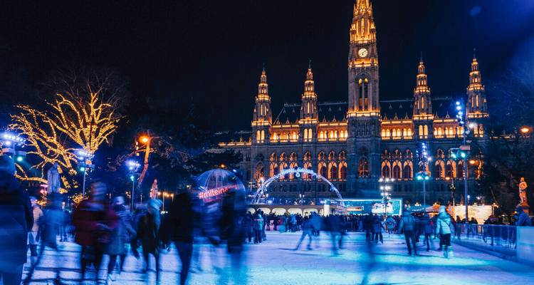Escena nocturna de patinaje sobre hielo frente al Rathaus de Viena iluminado; patinadores desenfocados en movimiento.