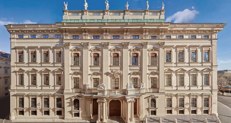 Fachada frontal de un ornado palacio barroco con estatuas en el techo contra un cielo azul brillante.