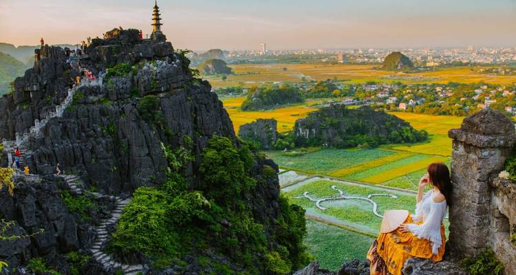 Un viajero en traje tradicional se sienta en una saliente rocosa con vista a los espectaculares acantilados de karst y arrozales en Ninh Binh al atardecer.