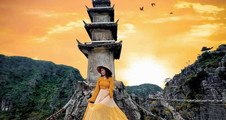 Una mujer en amarillo posa frente a una pagoda de piedra con un cielo naranja vivido sobre acantilados de piedra caliza.