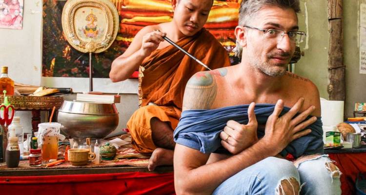A monk applies a traditional tattoo to a seated traveler inside a small studio filled with ritual objects.