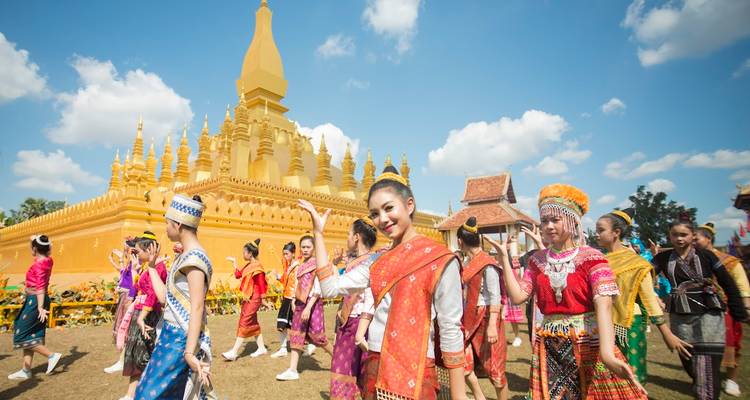 Les danseurs vêtus de costumes traditionnels laotiens colorés défilent devant la stupa dorée de Pha That Luang sous un ciel bleu.