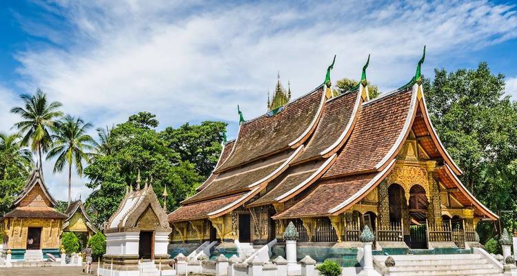 Temple Wat Xieng Thong aux ornements en bois finement travaillés, encadré par des palmiers et un ciel d'un bleu éclatant.