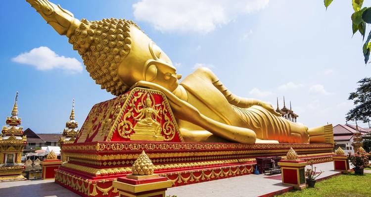 Une massive statue de Bouddha doré allongé repose sur un piédestal richement orné sous un ciel dégagé.