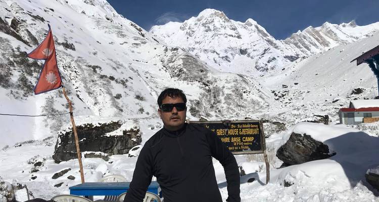 Trekker met zonnebril staat naast het bord van Annapurna Base Camp, omringd door diepe sneeuw en torenhoge bergtoppen.