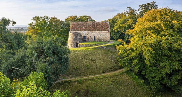 Historische Steinkapelle auf einem grasbewachsenen Bergrücken, umrahmt von üppiger Vegetation im frühen Morgenlicht