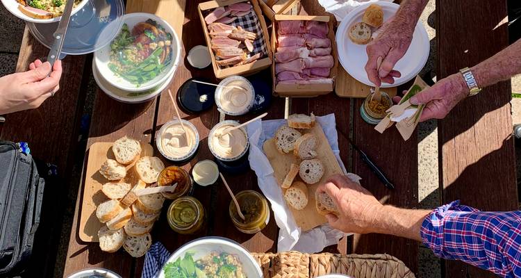 Picnic rústico al aire libre con panes, carnes, ensaladas, quesos y frascos de condimentos siendo compartidos.