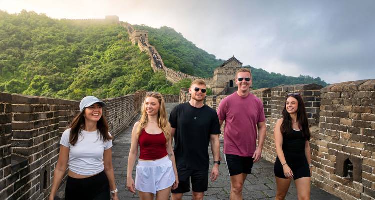 Eine fröhliche Gruppe spaziert entlang eines antiken Steinabschnitts der Chinesischen Mauer mit grünen Hügeln, die sich dahinter erstrecken.