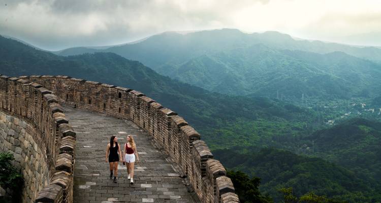 Zwei Frauen spazieren einen gewundenen Abschnitt der Chinesischen Mauer entlang, die über nebelverhangene grüne Berge blickt.