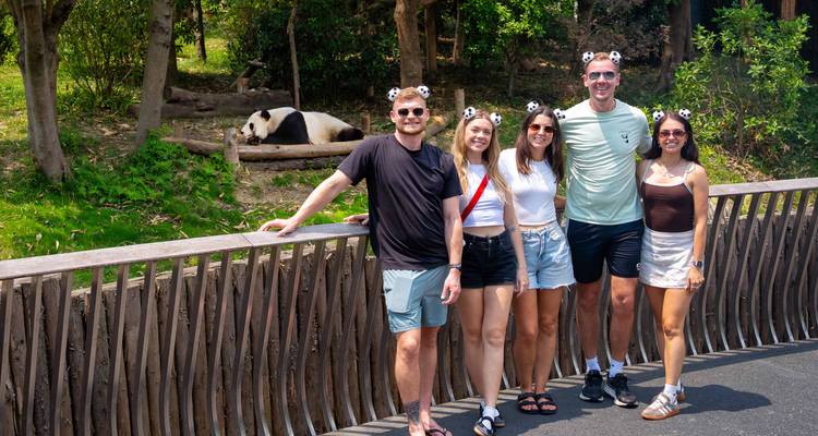 Besucher mit Pandaohren posieren neben einem ruhenden Panda in einem mit Blättern bewachsenen Gehege.