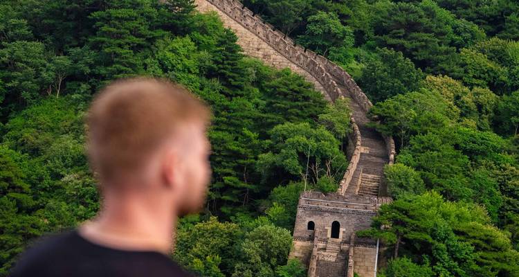 Ein Reisender blickt auf einen fernen, sich windenden Abschnitt der Chinesischen Mauer, der in einen dichten grünen Wald verschwindet.