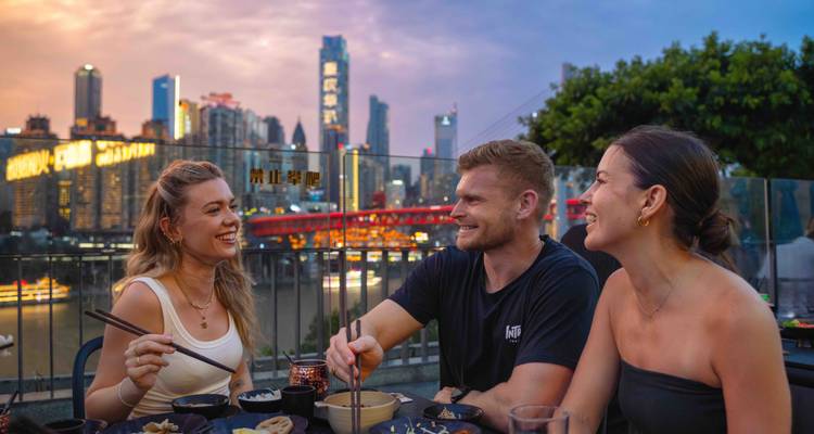 Freunde genießen die Freiluftküche in der Dämmerung mit der Skyline von Chongqing und einer beleuchteten Brücke im Hintergrund.