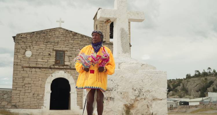 Homme tarahumaras vêtu de vêtements traditionnels aux couleurs vives se tient à côté d'une croix en pierre blanche devant une ancienne église missionnaire