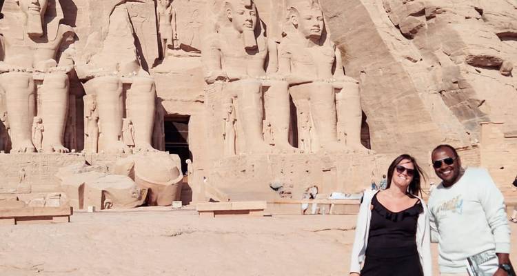 Un couple souriant se tient devant les statues monumentales d'Abu Simbel dans la lumière éclatante du désert.