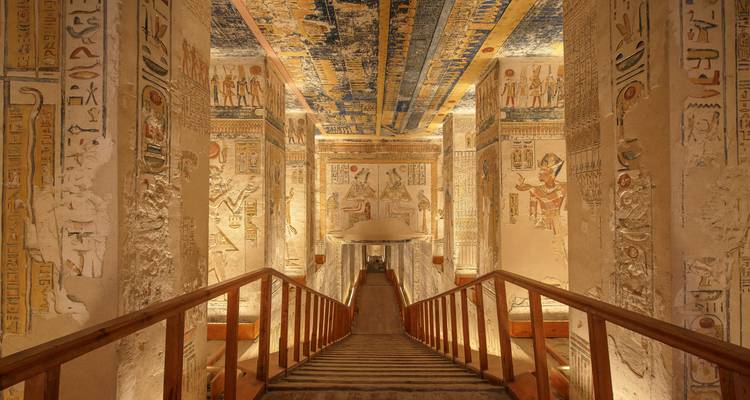 Intérieur d'une ancienne tombe égyptienne illuminée avec des colonnes richement peintes et un escalier en bois.