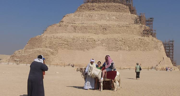 Des visiteurs à dos de chameau posant devant la Pyramide à degrés de Saqqara lors d'une journée désertique dégagée.