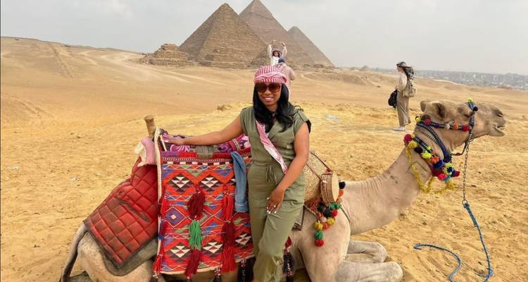Un viajero posando con un camello decorado frente a las pirámides de Giza en una mañana desértica brumosa.
