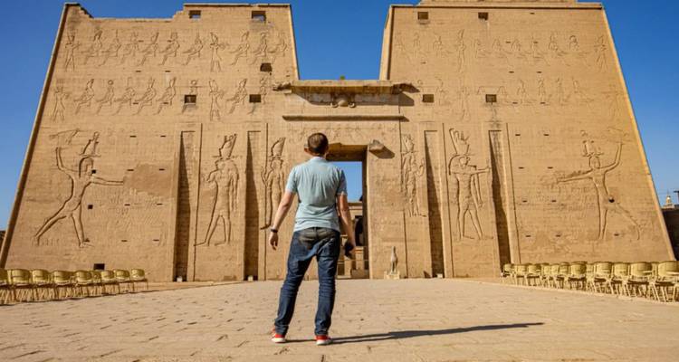 El viajero está frente a los imponentes pilonos del Templo de Edfu decorados con relieves del antiguo Egipto bajo un cielo azul profundo.