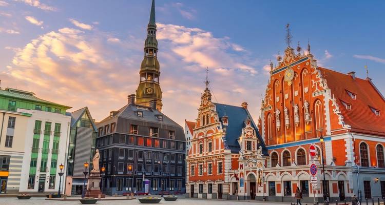 Edificios medievales coloridos y la alta aguja de la plaza del Casco Antiguo de Riga bajo nubes vespertinas en tonos pastel.