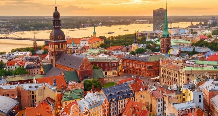 Puesta de sol dorada sobre el horizonte de Riga, con el río Daugava y la torre de la catedral histórica visible desde arriba.