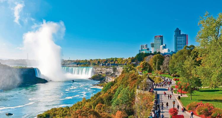 Malerischer Fußweg entlang des Niagara River mit Stadtskyline und Gischt der Fälle, die in der Sonne aufsteigt