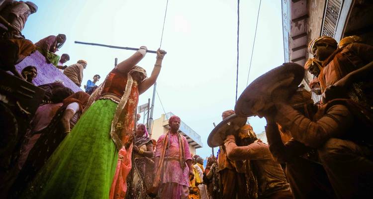 Femme en jupe verte qui agite avec enjouement un bâton au-dessus d'hommes portant des boucliers lors des rituels de rue du Holi Lathmar.
