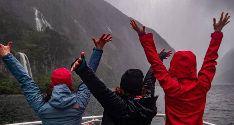 Trois voyageurs réunis en groupe sur un bateau lèvent les bras avec joie vers les chutes d'eau brumeuses du Milford Sound.