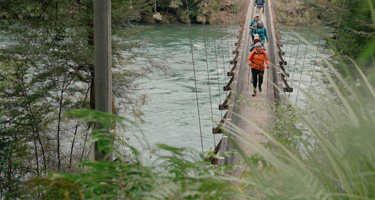 Une file de randonneurs traversant un pont suspendu étroit au-dessus d'une rivière au cours rapide entourée de forêt.
