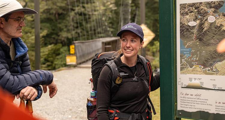 Un guide souriant avec un sac à dos explique une carte de sentier à un voyageur au point de départ d'un sentier forestier.