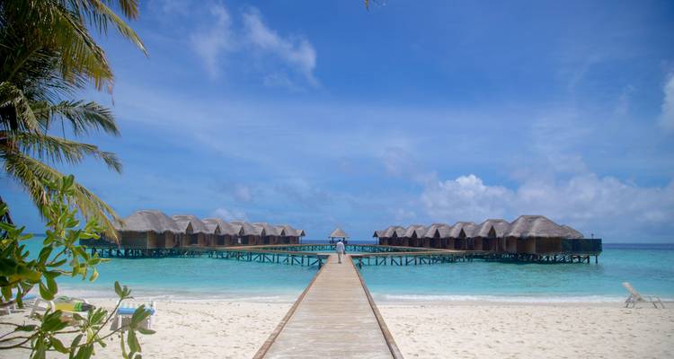 Passerelle en bois menant à une rangée de villas sur pilotis au-dessus d'une mer turquoise et d'une plage de sable blanc.