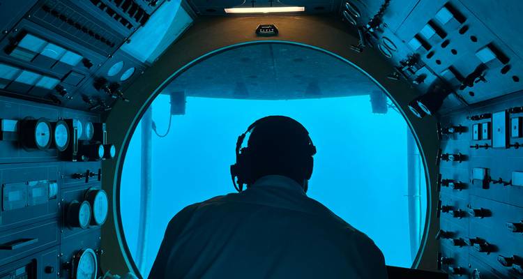 Opérateur en silhouette portant un casque audio à l'intérieur d'un petit sous-marin, regardant par une grande fenêtre circulaire vers l'eau bleue.