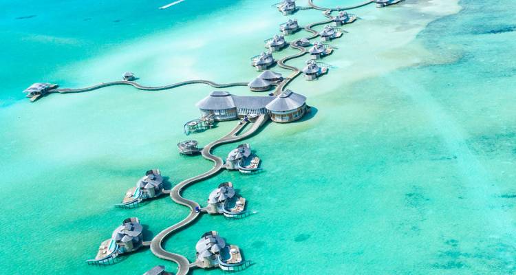 Vue aérienne de villas de luxe surplombant l'eau reliées par des passerelles en bois sinueuses dans un lagon d'eau cristalline bleu azur.