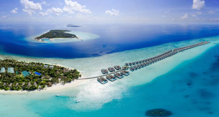 Vaste panorama aérien d'une station balnéaire insulaire avec des rangées de bungalows sur l'eau et une mer bleu profond s'étendant jusqu'à l'horizon.