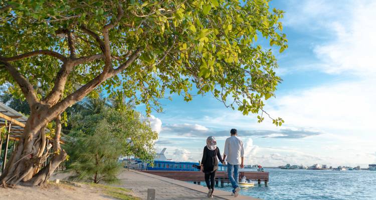 Un couple se promène main dans la main le long d'une promenade en bord de mer ombragée par des arbres feuillus avec la mer bleue au-delà.