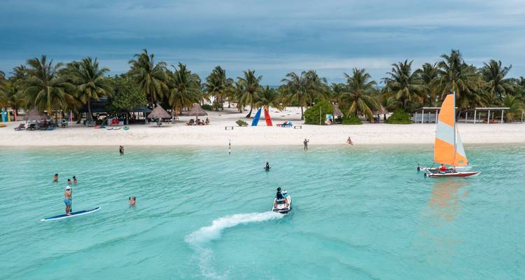 Scène de lagon avec des personnes pratiquant le paddle-board, le jet-ski et la voile devant une plage de sable bordée de palmiers.