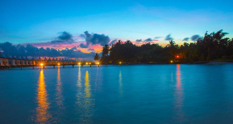 Le ciel du coucher de soleil aux couleurs vives se reflète dans le lagon tranquille avec des bungalows sur pilotis illuminés par des lumières dorées.
