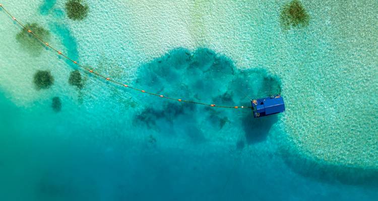 Vue aérienne par drone d'un lagon turquoise avec une petite cabane flottante bleue maintenue par une corde au-dessus du fond marin sablonneux.
