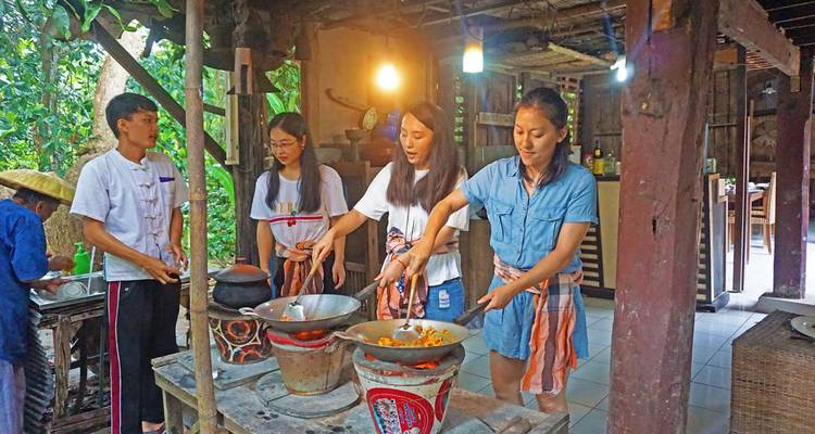 Los viajeros revuelven woks en una cocina rústica durante una clase de cocina tailandesa iluminada por bombillas cálidas.
