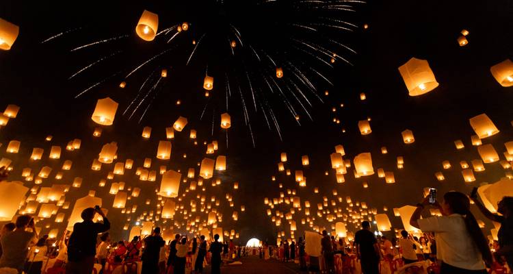 Des centaines de lanternes en papier luminescentes s'élèvent dans un ciel sombre avec des feux d'artifice alors que de grandes foules se rassemblent lors d'un festival thaïlandais.