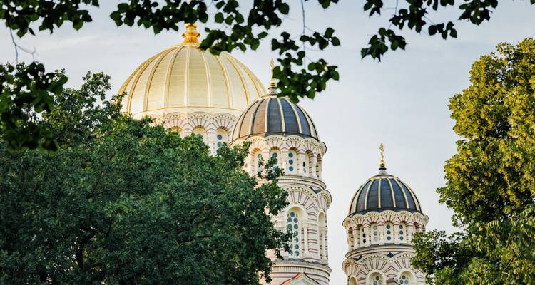 Las cúpulas doradas y torres ornamentadas de la Catedral de la Natividad de Riga enmarcadas por ramas frondosas.