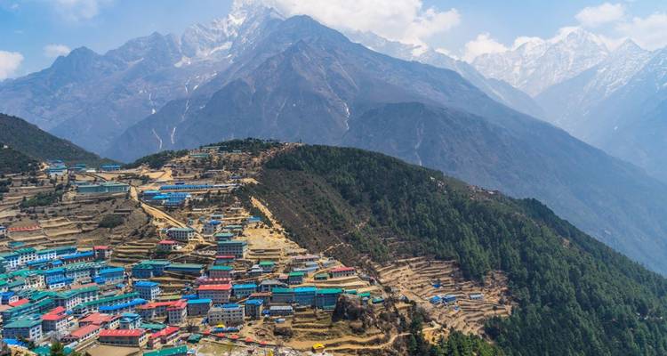 Les toits colorés du Bazar de Namche regroupés sur une crête forestière escarpée avec l'imposante chaîne de l'Himalaya en arrière-plan.