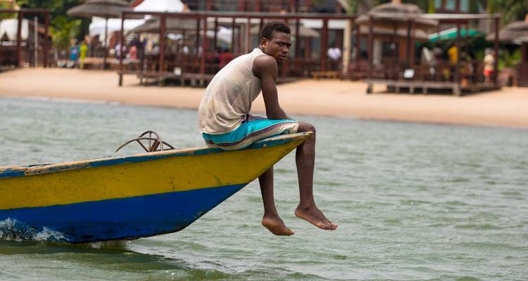Un jeune homme est assis à la proue d'un bateau de pêche en bois peint qui flotte au large d'une plage tropicale de sable fin.